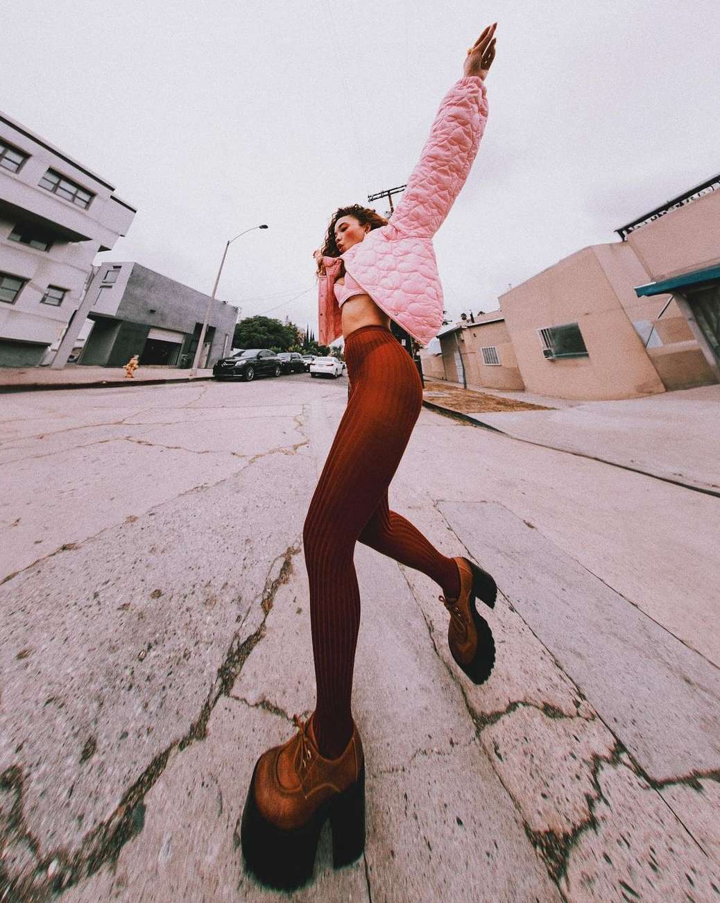 A young adult woman in a pink jacket and brown platform shoes is seen on a cracked asphalt street under a cloudy sky in an urban area.