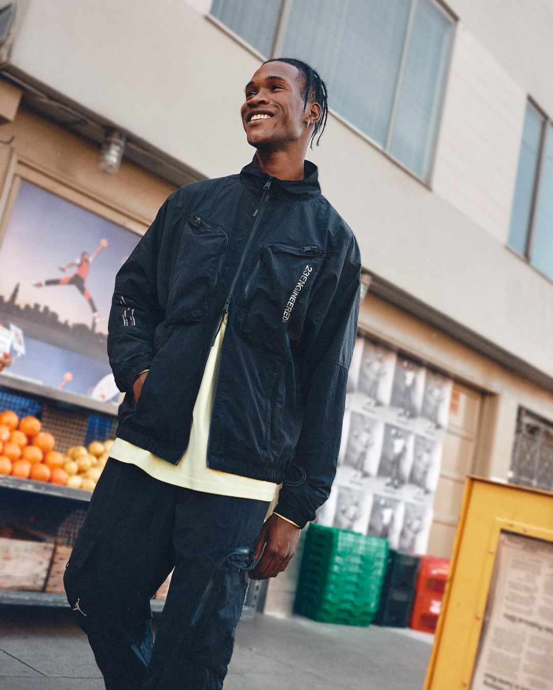 Stylish man smiles outside in black jacket and pants near a building with oranges and a poster visible in the background.