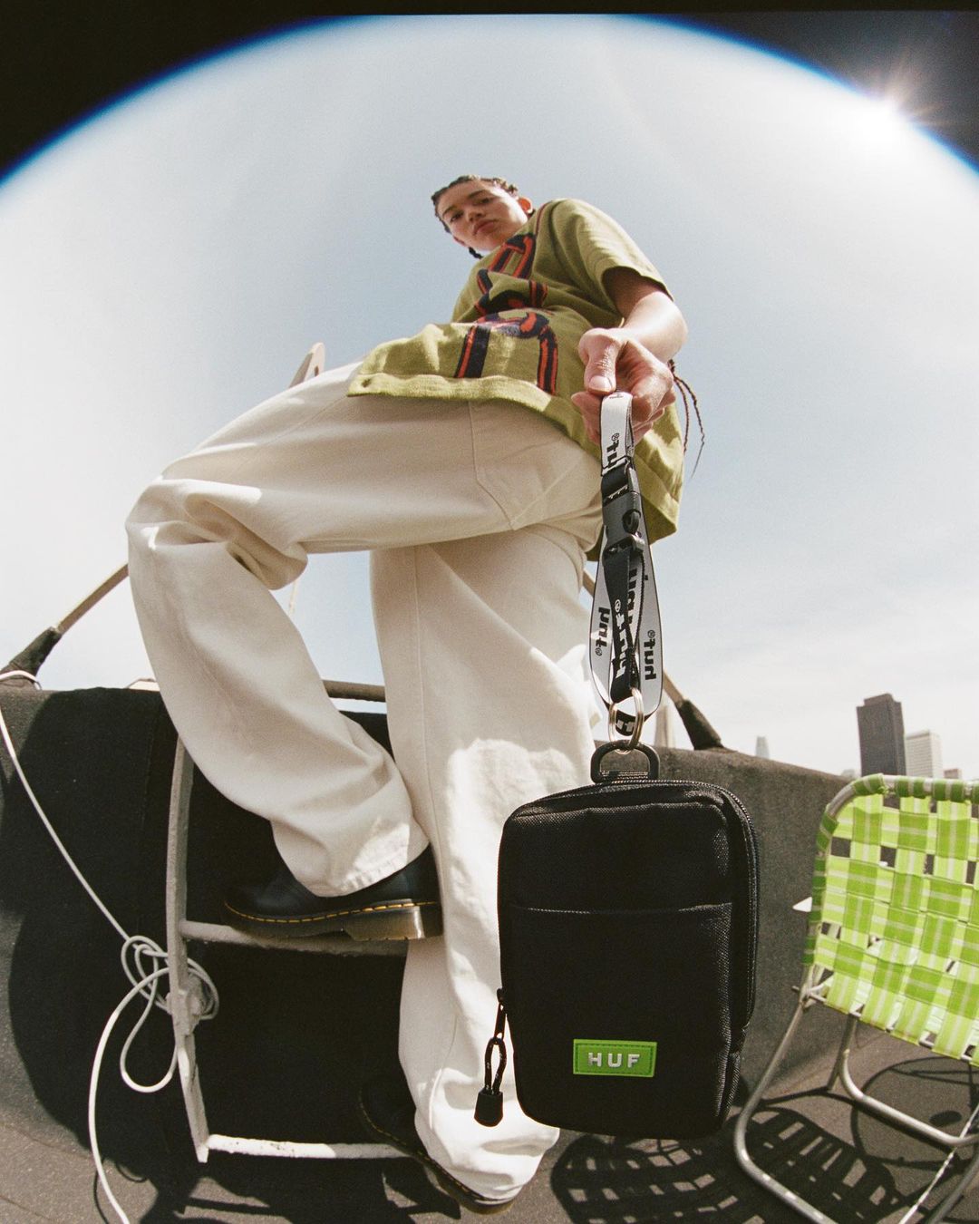 A young adult poses on a rooftop in an eye-level shot, wearing casual clothing and holding a black HUF pouch in an outdoor setting.