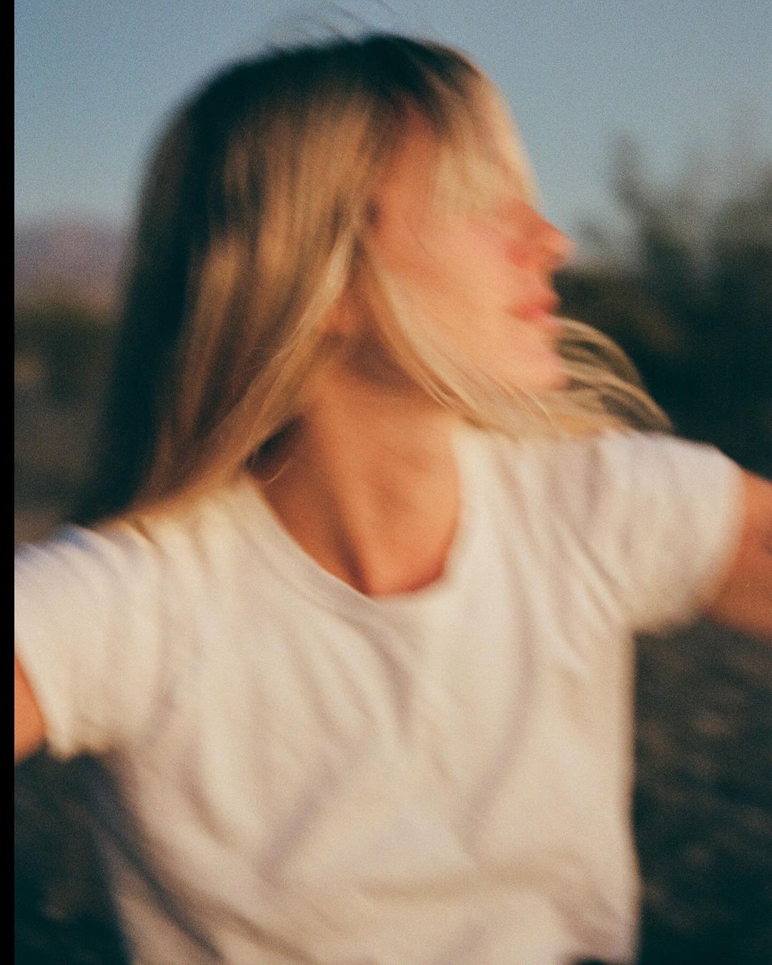 Blurred image of a blonde woman with a white t-shirt outdoors in a desert environment with mountains in the distance and a soft, sunny light.