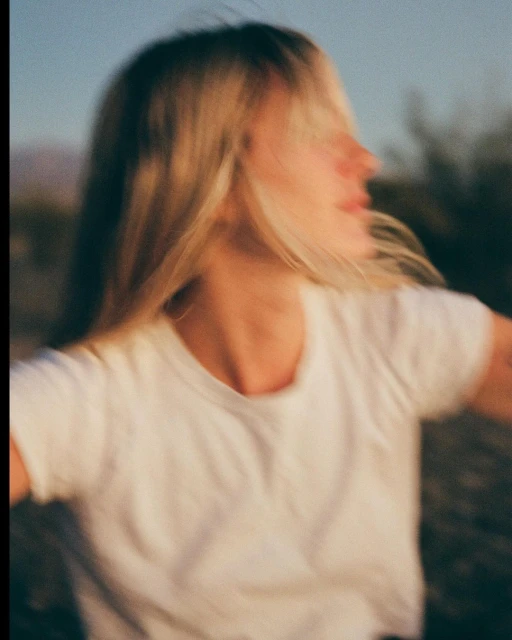 Blurred image of a blonde woman with a white t-shirt outdoors in a desert environment with mountains in the distance and a soft, sunny light.