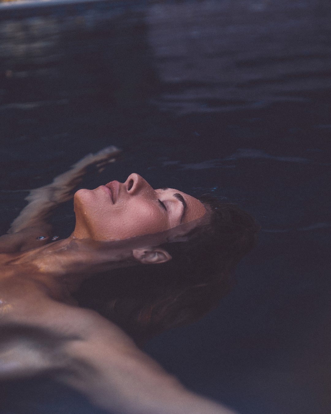 Woman floats serenely in dark water, face up, eyes closed, showcasing an intimate and tranquil moment of relaxation in a pool.