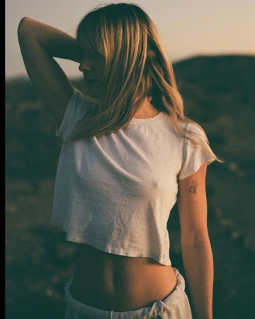 Woman with blonde hair wearing a white t-shirt and grey trousers posing outdoors in natural light.