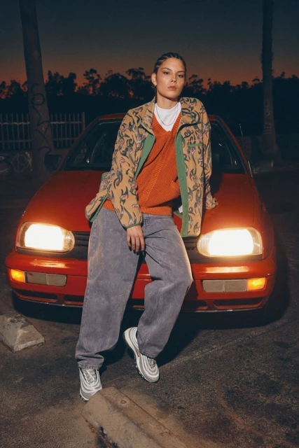 Stylish young woman in a patterned jacket leans on a red car at dusk, showcasing casual fashion in an urban setting with soft lighting.
