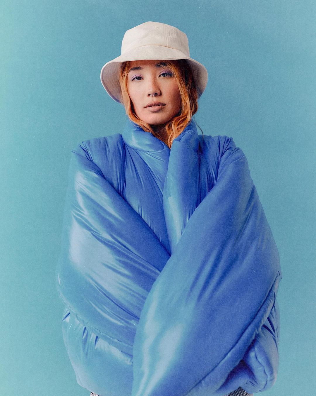 Portrait of a woman wearing a blue puffer jacket and a white bucket hat against a blue backdrop by photographer Arturo Torres.