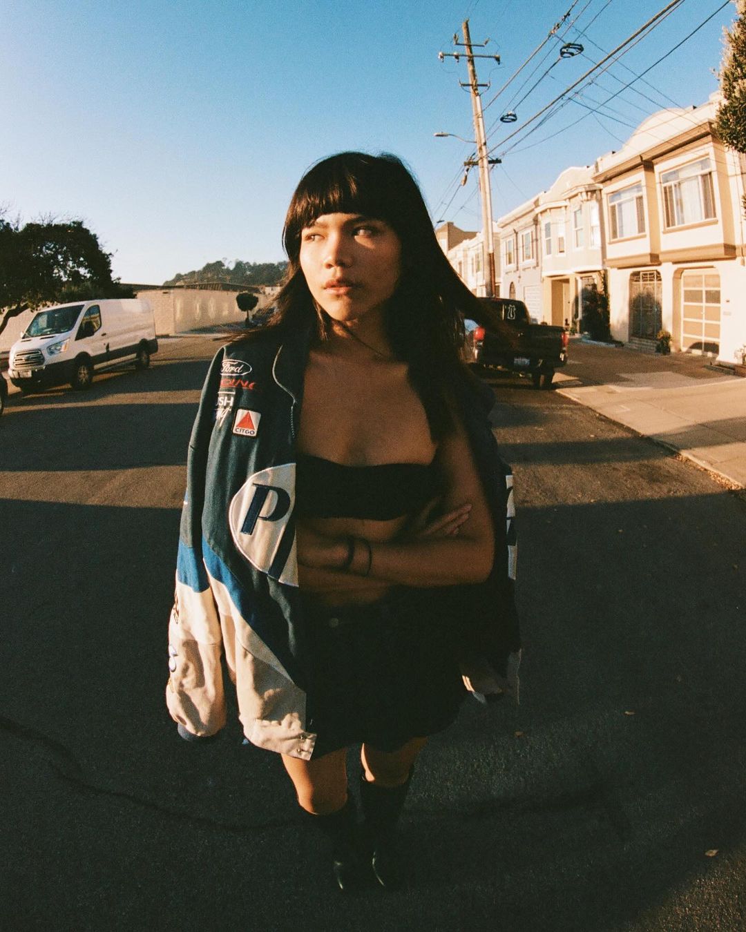 Young woman in an oversized jacket stands on a city street during the day with residential buildings and vehicles in the background.