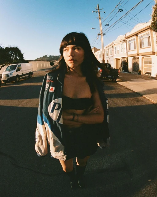 Young woman in an oversized jacket stands on a city street during the day with residential buildings and vehicles in the background.