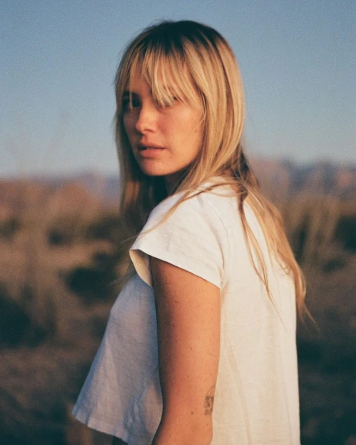 A young adult woman with blond hair and bangs looks over her shoulder outdoors with a neutral facial expression in a landscape setting.