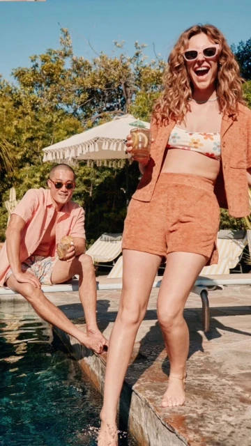 Two adults near a pool on a sunny day. A woman in floral swimwear and peach suit holds a drink. A man sits with a coconut.
