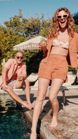 Two adults near a pool on a sunny day. A woman in floral swimwear and peach suit holds a drink. A man sits with a coconut.