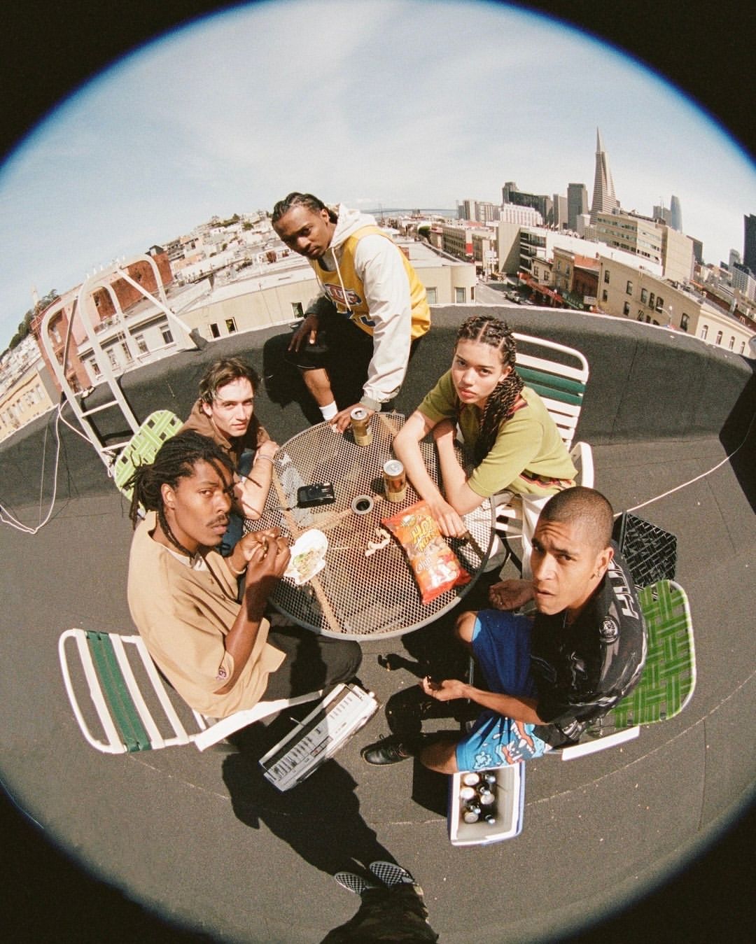 Group of six young adults gathered around a table on a rooftop with an urban skyline, enjoying snacks and drinks on a sunny day.
