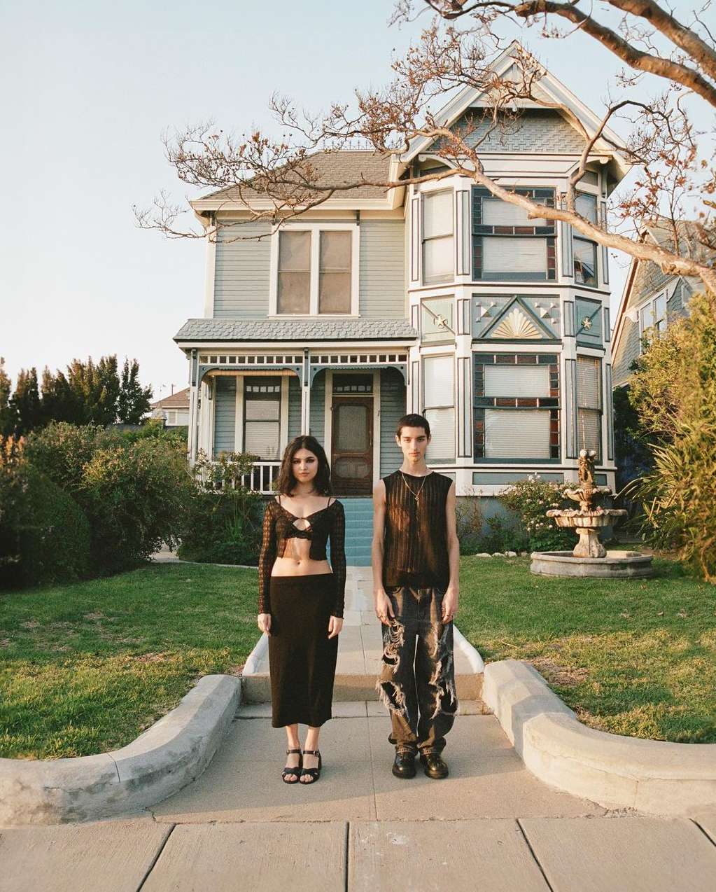 Two adults standing in front of a two-story blue house with a well-kept lawn and sidewalk on a sunny day.