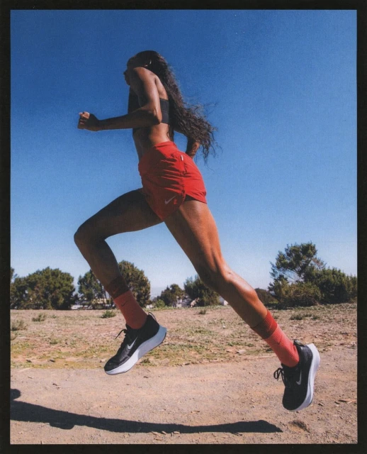 Athletic woman in running attire runs outdoors on a dirt trail with sparse trees under a clear blue sky in a medium, full shot.