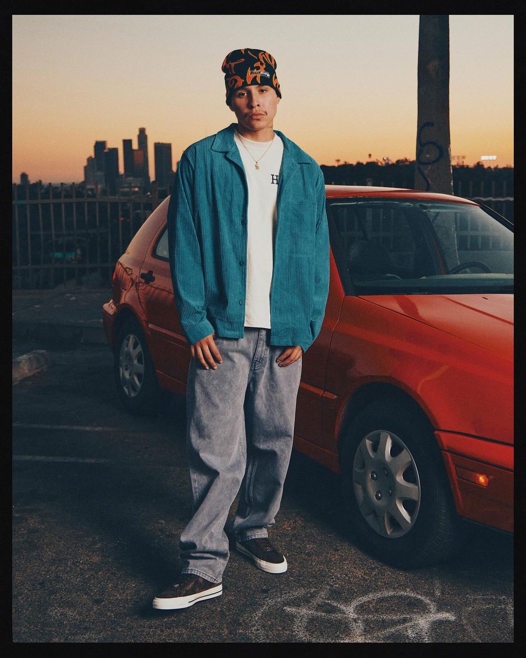 An adult male in a beanie, turquoise jacket, and grey jeans poses next to a red car in an urban setting at sunset, with city skyline backdrop.