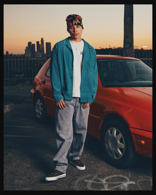 An adult male in a beanie, turquoise jacket, and grey jeans poses next to a red car in an urban setting at sunset, with city skyline backdrop.
