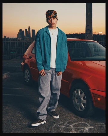 An adult male in a beanie, turquoise jacket, and grey jeans poses next to a red car in an urban setting at sunset, with city skyline backdrop.