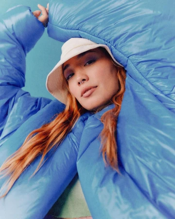 Portrait of a woman wearing a blue puffer jacket and a white bucket hat against a blue background