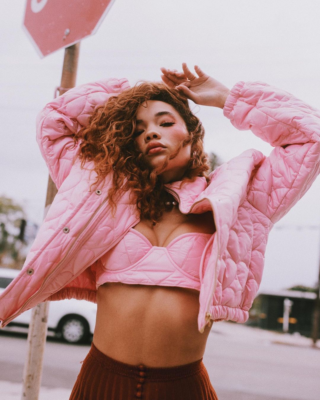 Young woman in pink quilted jacket and bra top poses outdoors near a stop sign, showcasing a fashion-forward, urban style.