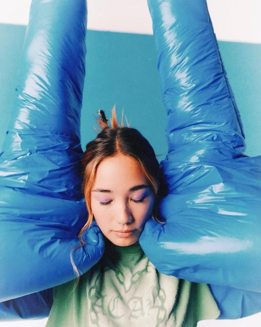 Young woman with light brown hair gazes downward with blue inflatable arms framing her face against a light blue background in portrait.