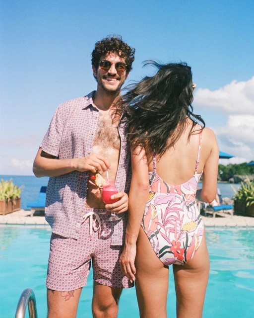 A couple stands by an outdoor pool on a sunny day. The man in a patterned shirt holds a pink drink, and the woman wears a floral swimsuit.