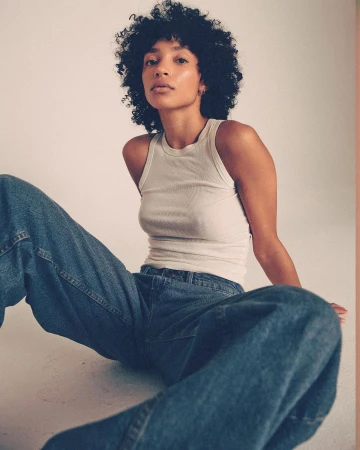 Woman with curly dark hair wearing a white tank top and blue denim jeans seated against a neutral studio backdrop