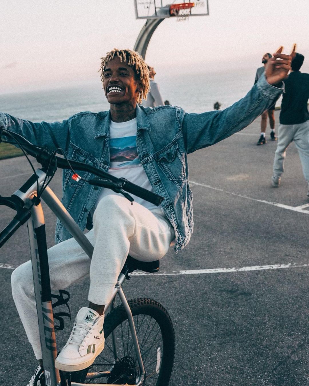 Smiling Black man with blond dreads rides a bike on an asphalt court near the ocean, wearing denim jacket and white sneakers, daytime.
