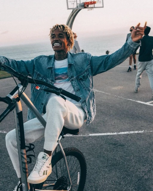 Smiling Black man with blond dreads rides a bike on an asphalt court near the ocean, wearing denim jacket and white sneakers, daytime.