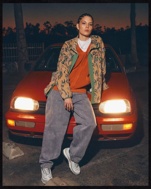 Stylish woman poses against a red car wearing patterned jacket, orange sweater, grey jeans, and sneakers in an outdoor setting at dusk.
