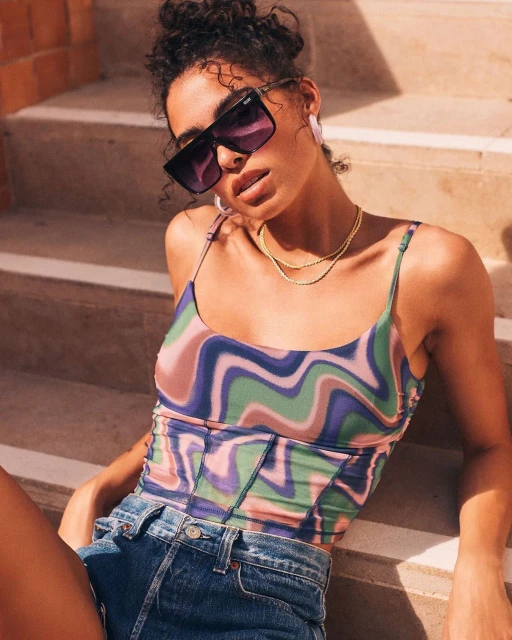 A young woman sits on outdoor steps wearing a retro-print top, sunglasses, and gold necklaces in a lifestyle fashion shot.