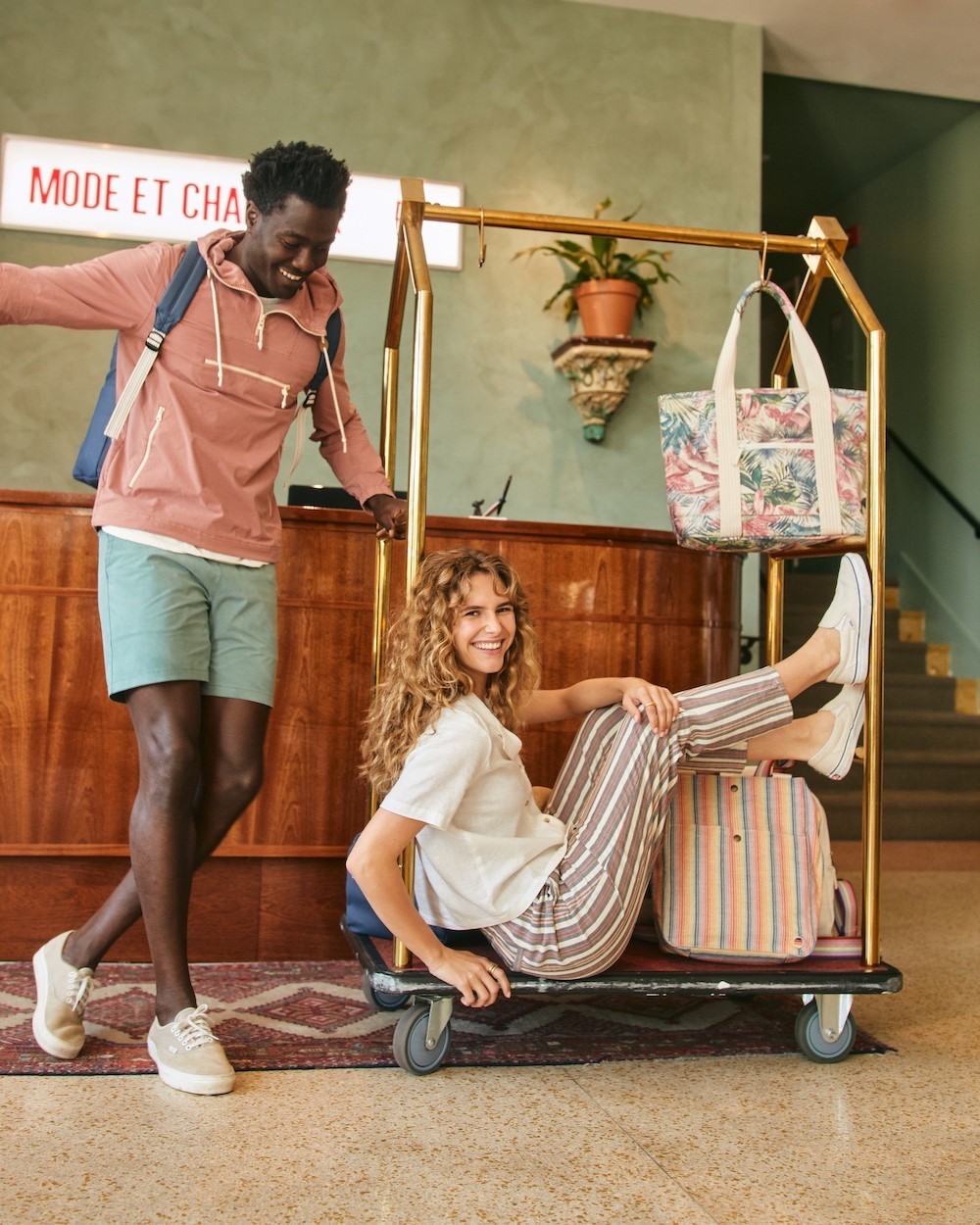 Happy diverse couple at hotel. African American man stands near luggage trolley as curly-haired woman sits among bags. Travel, tourism.