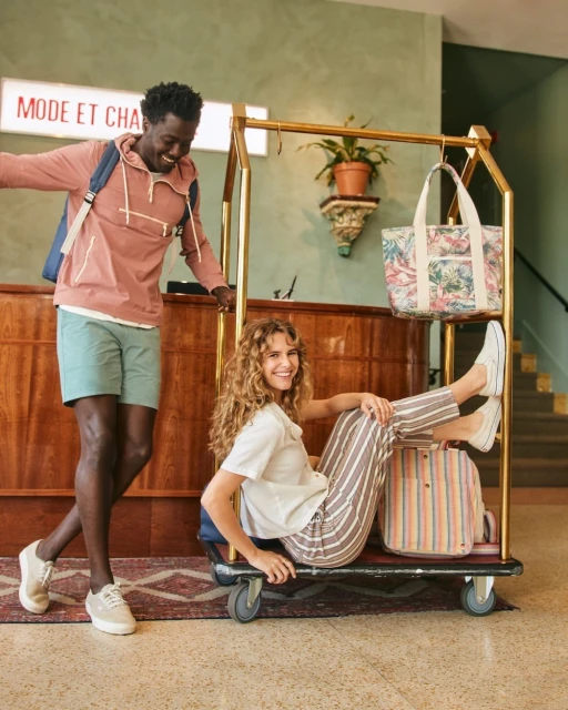 Happy diverse couple at hotel. African American man stands near luggage trolley as curly-haired woman sits among bags. Travel, tourism.