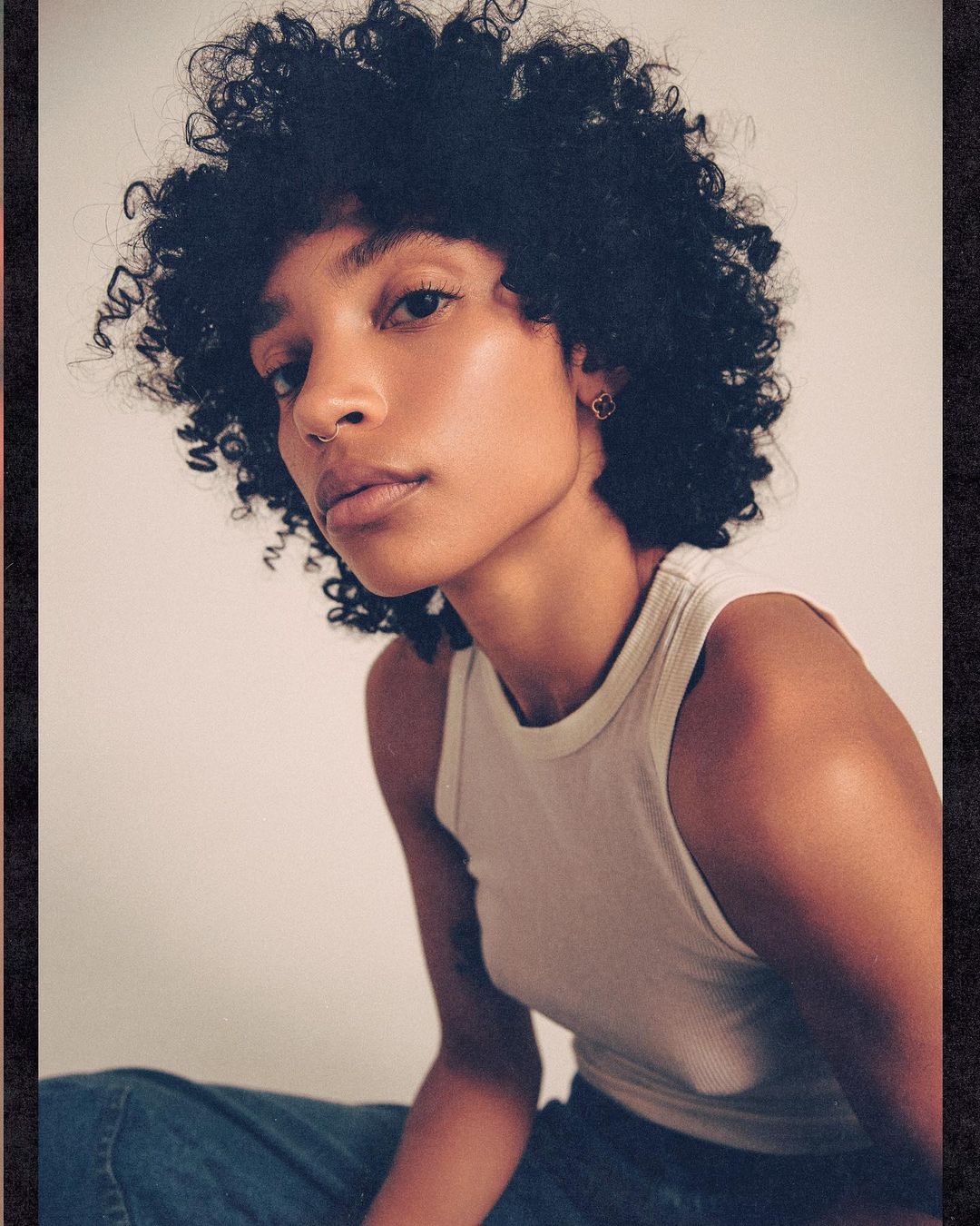 Portrait of a young adult woman with dark curly hair and a neutral expression, wearing a tank top and jeans against a white background.