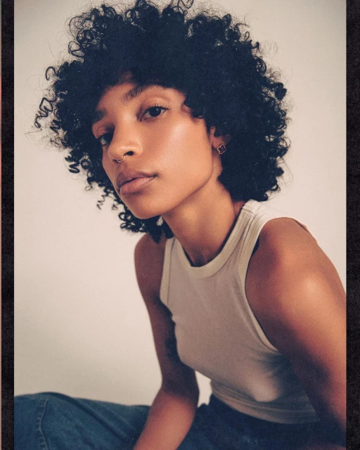 Portrait of a young adult woman with dark curly hair and a neutral expression, wearing a tank top and jeans against a white background.