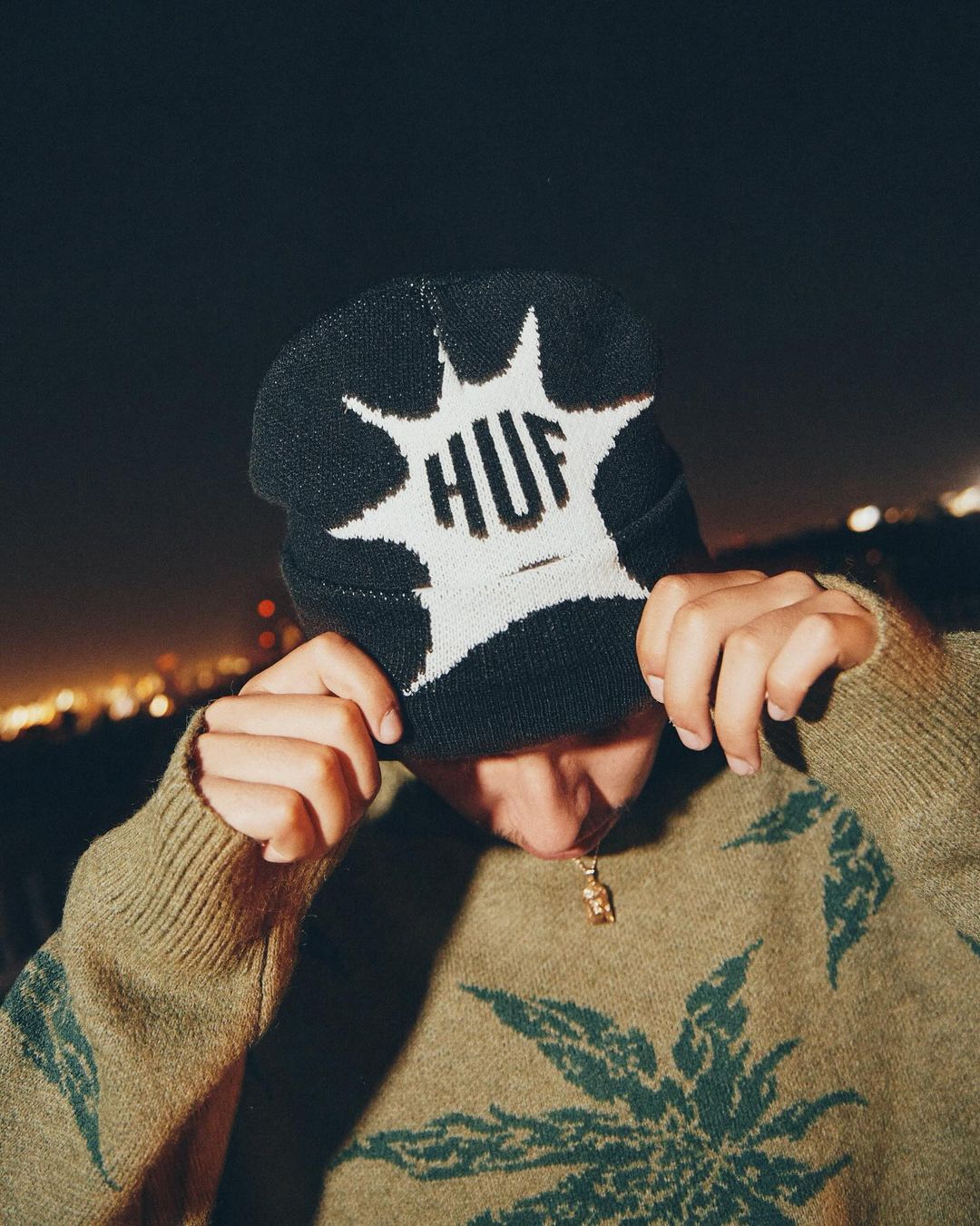 Person in HUF beanie and sweater with cannabis leaf design against blurred city night lights, shot at eye-level.