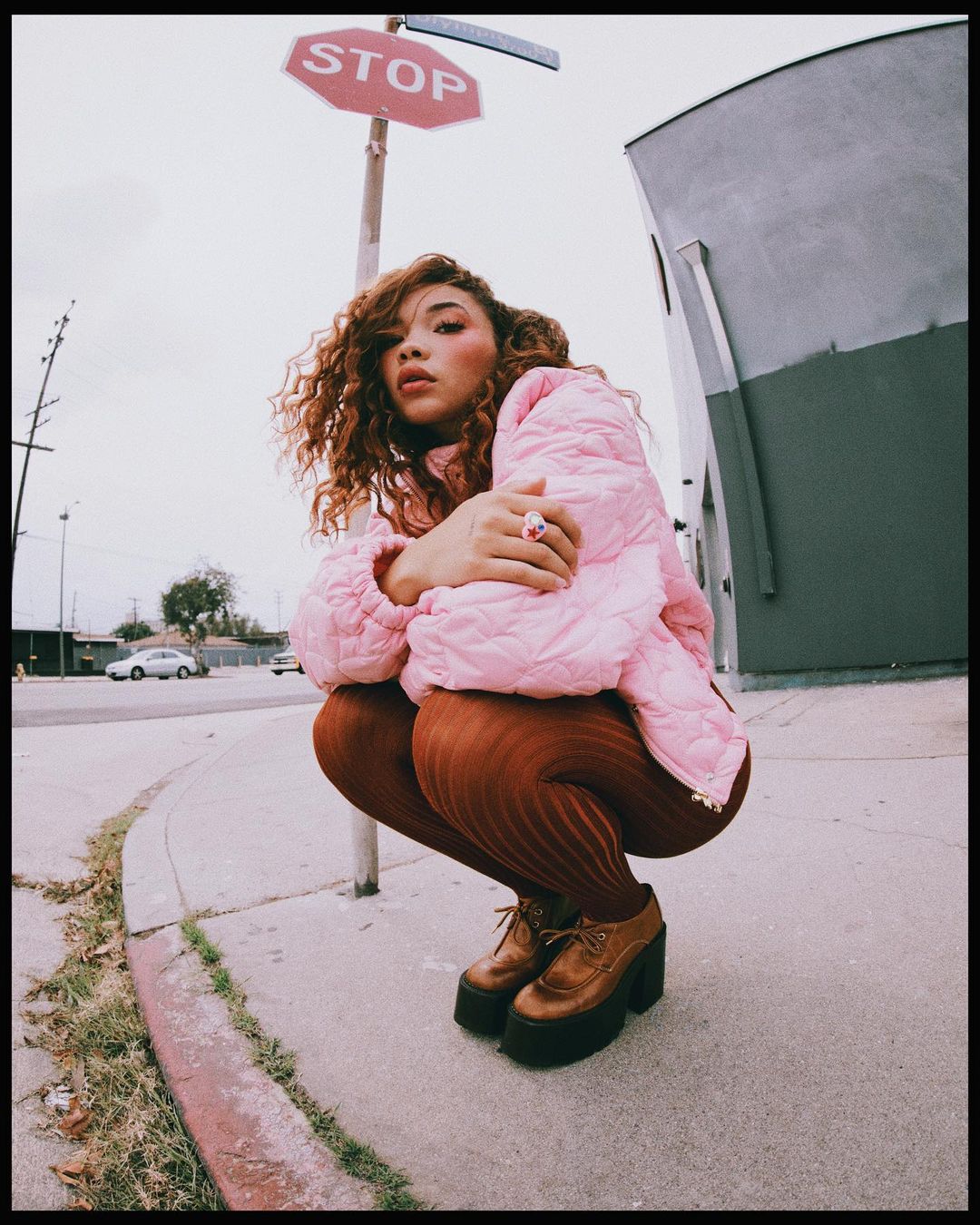 Woman with curly hair wearing a pink puffer jacket crouching near a red stop sign on an overcast day in an urban environment.