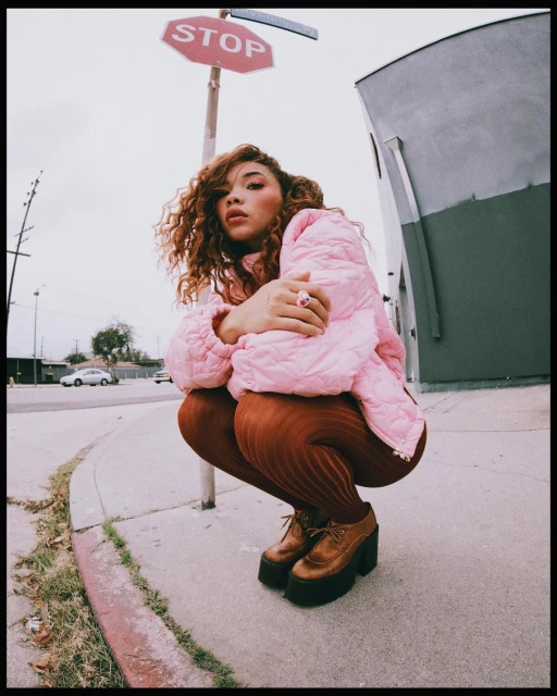 Woman with curly hair wearing a pink puffer jacket crouching near a red stop sign on an overcast day in an urban environment.