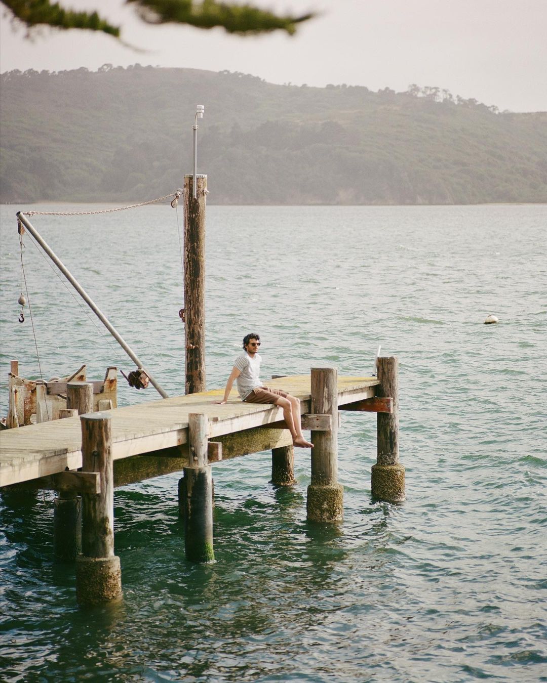 Man sits on a wooden dock over calm water with green tree-covered mountains in the background, enjoying a serene outdoor moment.