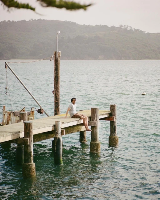 Man sits on a wooden dock over calm water with green tree-covered mountains in the background, enjoying a serene outdoor moment.