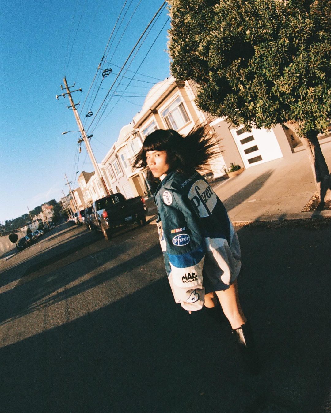 Model with dark hair wearing a blue racing jacket walking down a sunny residential street photographed by Arturo Torres.