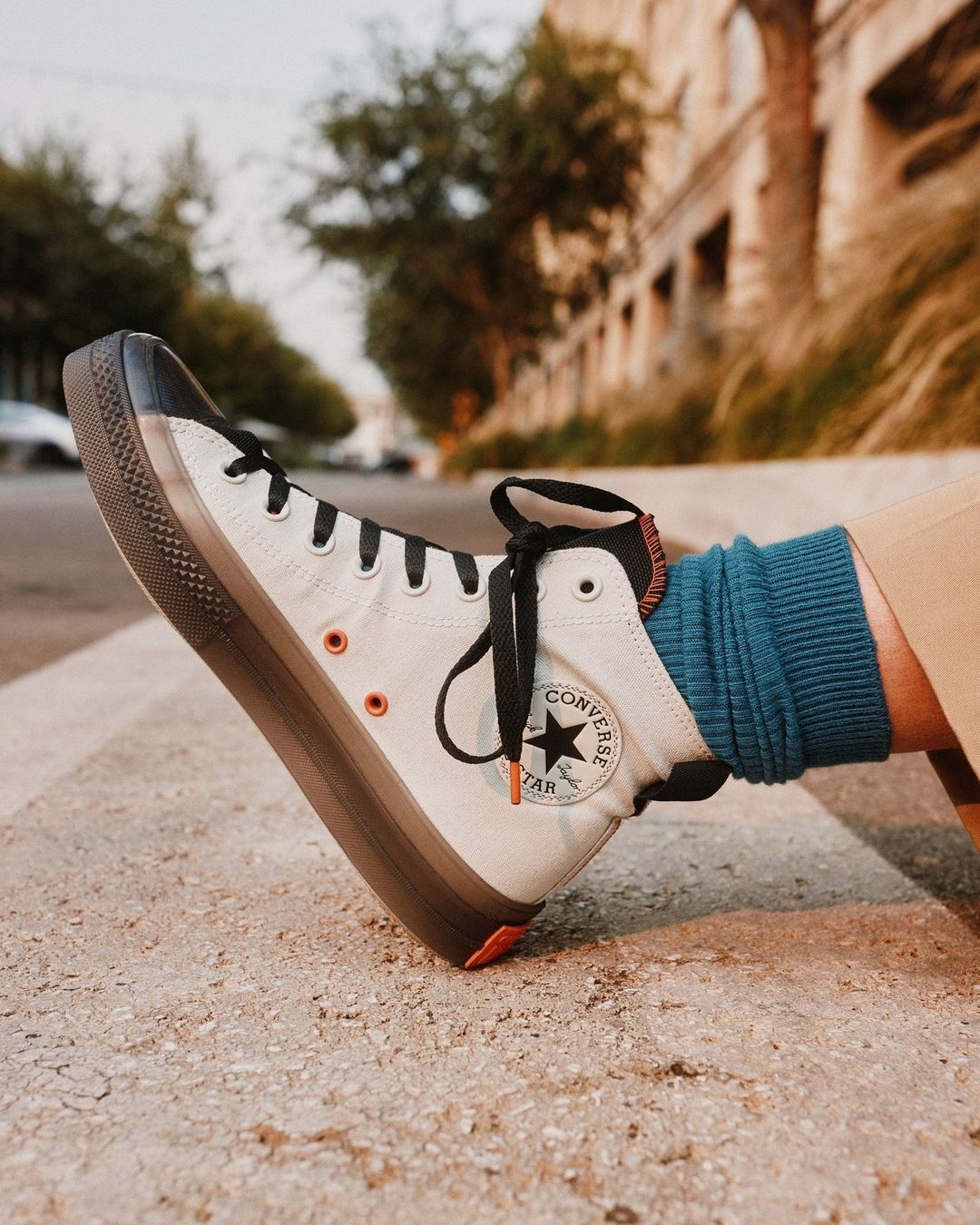 Close-up of a white Converse high-top sneaker with a thick brown sole, worn with blue socks, outdoors on concrete with building background.