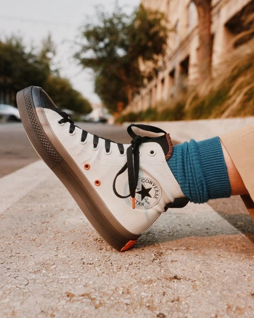 Close-up of a white Converse high-top sneaker with a thick brown sole, worn with blue socks, outdoors on concrete with building background.