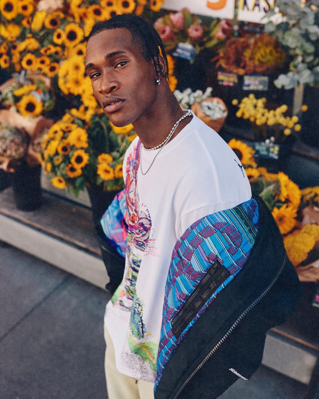 Portrait of a Black man wearing a patterned t-shirt and a black jacket standing in front of a flower stand with sunflowers.