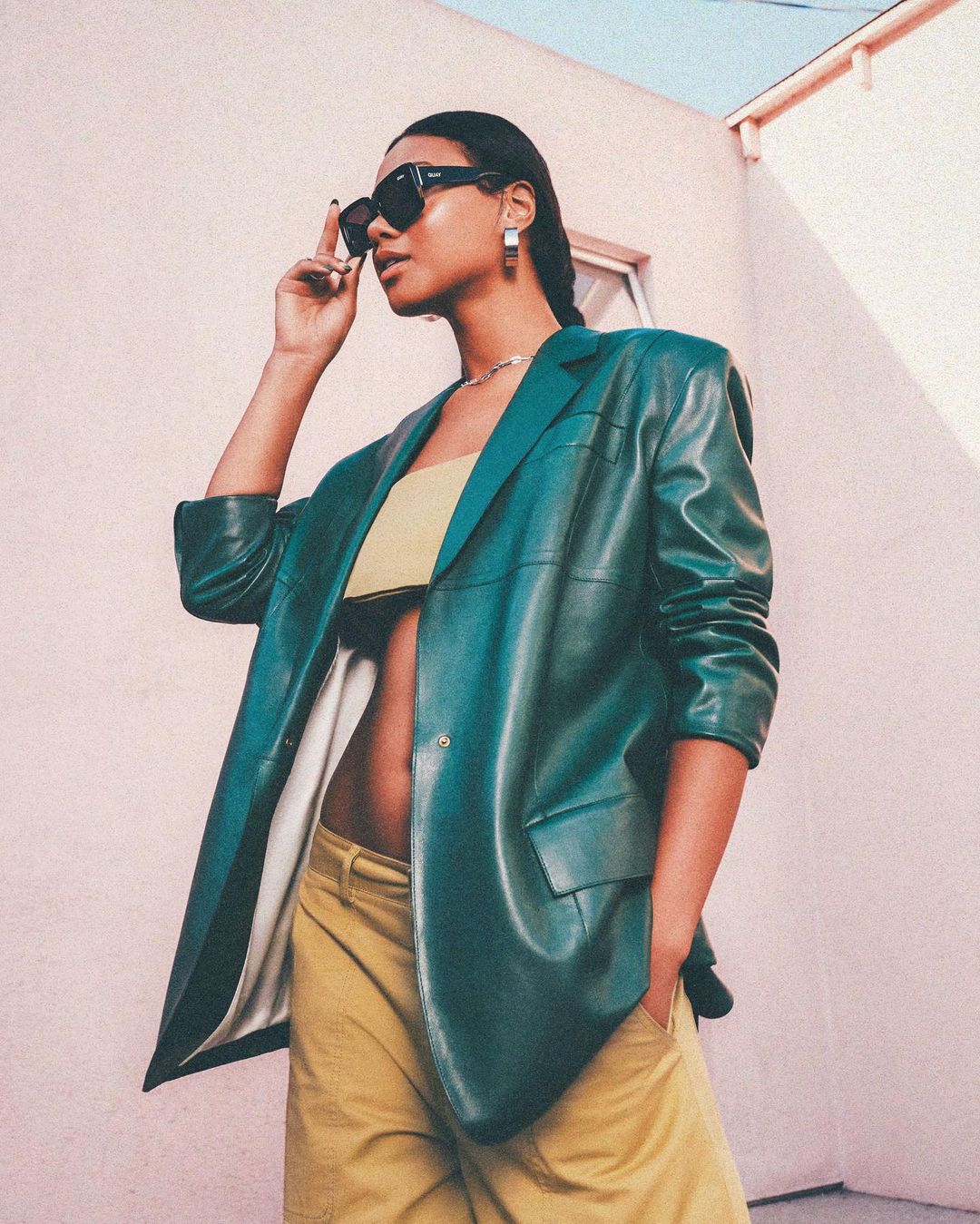 Woman in oversized green leather blazer and yellow pants poses outside, accessorized with sunglasses and hoop earrings in a fashion shot.