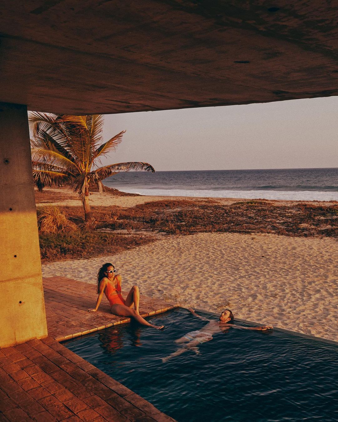 Two women relax in a poolside setting overlooking a beach with gentle waves, under a concrete structure, bathed in the warm light of dusk.