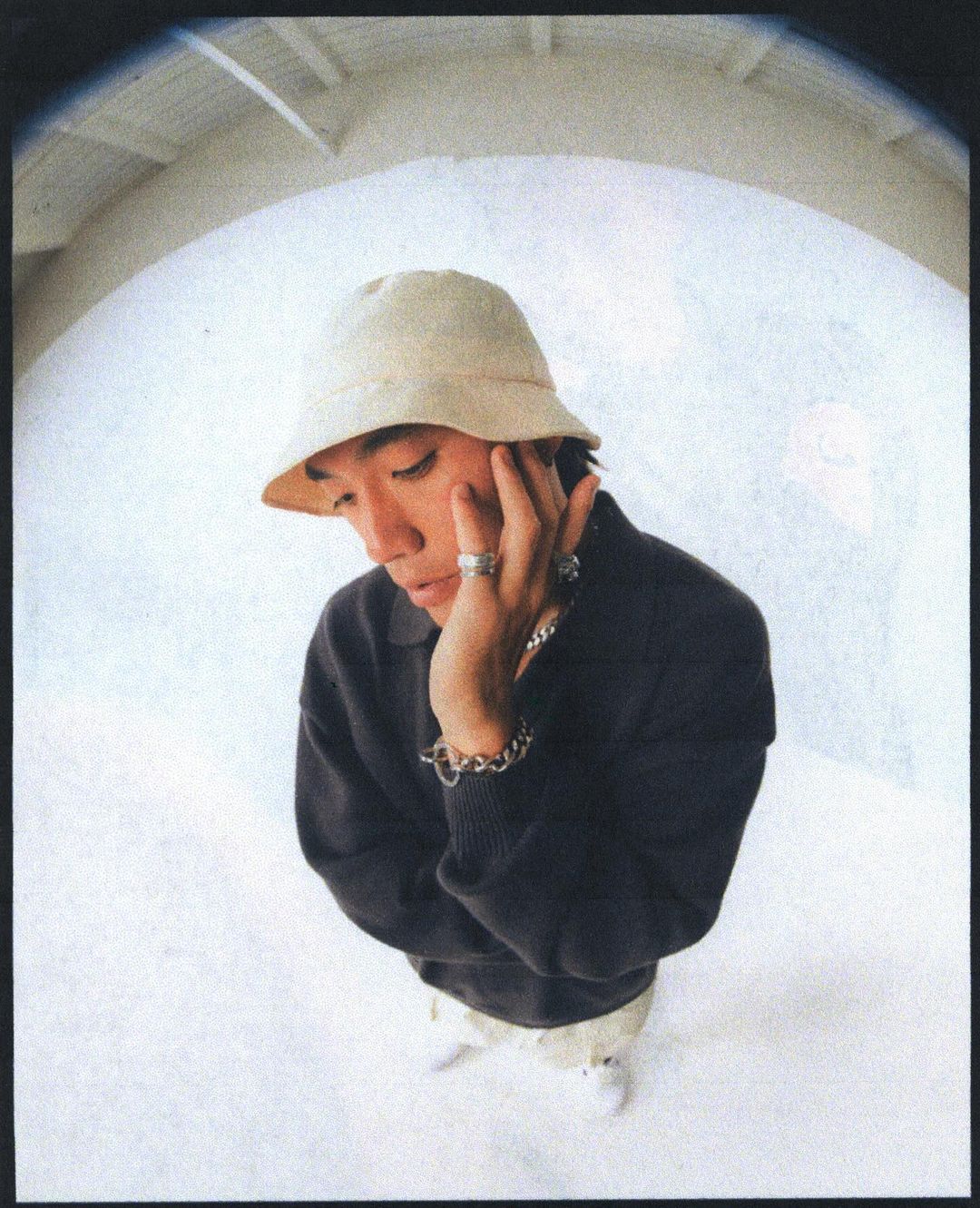 Portrait of a man wearing a bucket hat and dark sweater photographed with a fish-eye lens against a curved wall