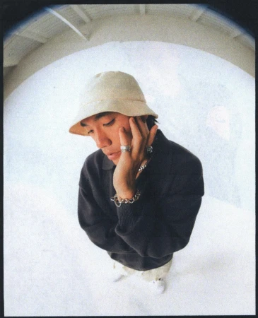 Portrait of a man wearing a bucket hat and dark sweater photographed with a fish-eye lens against a curved wall