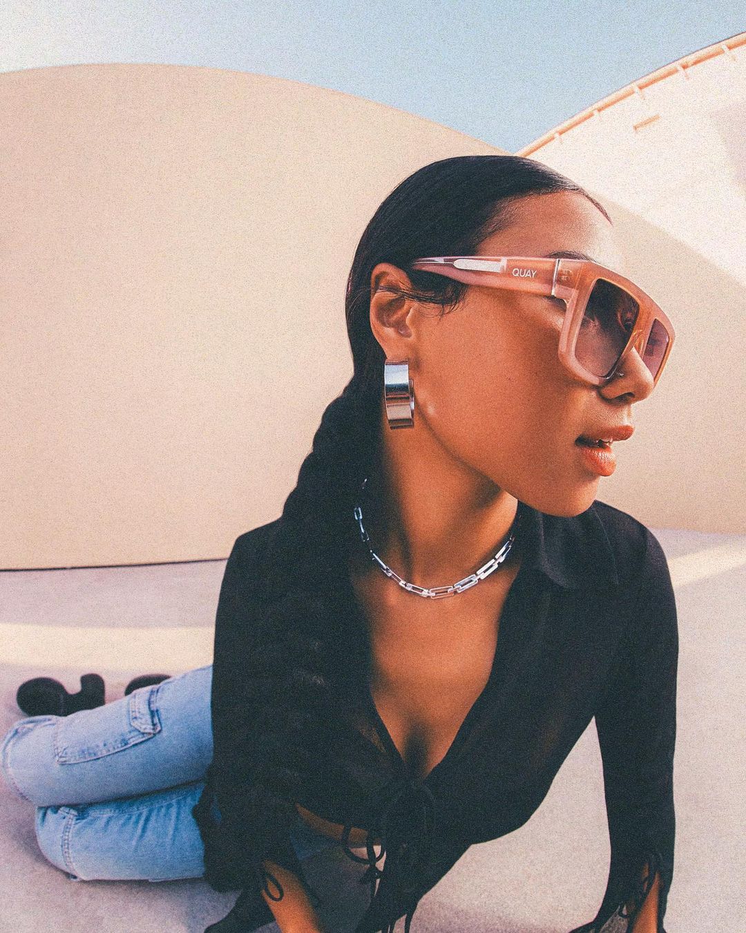 Woman with box-framed glasses and silver earrings and necklace, kneeling on the floor in sunlight, wearing a black top and light-blue jeans.