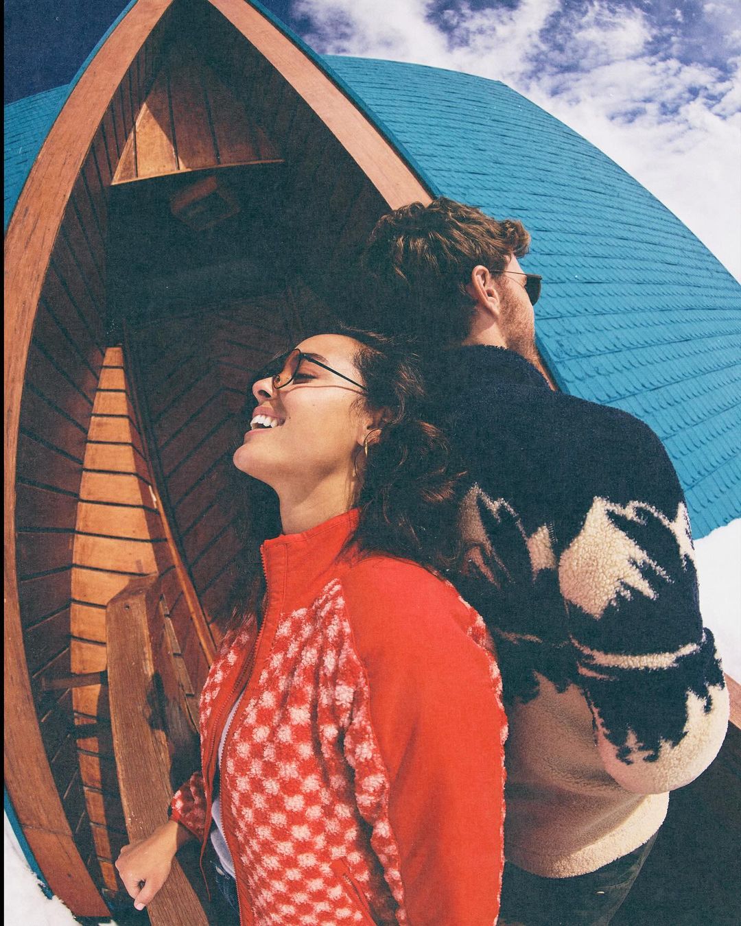 Young couple in front of a blue and wood building with a fisheye lens, woman smiling, man in sunglasses, blue sky with clouds above.