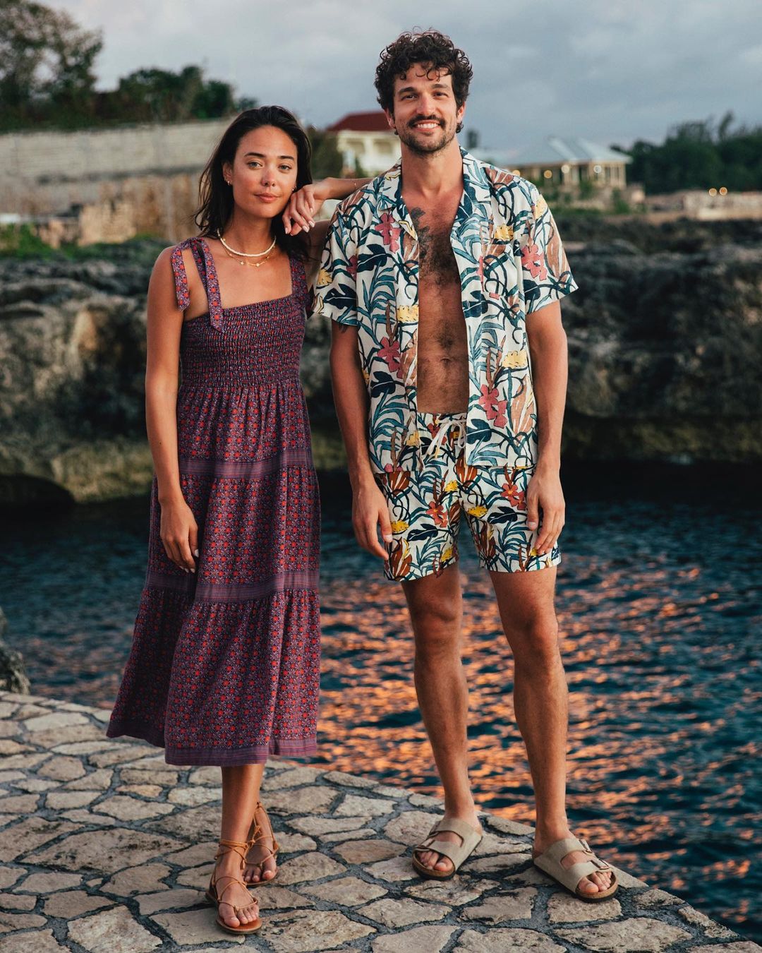 A happy couple poses in tropical resort wear near the ocean. The woman wears a floral sundress; the man, a matching shirt and shorts.