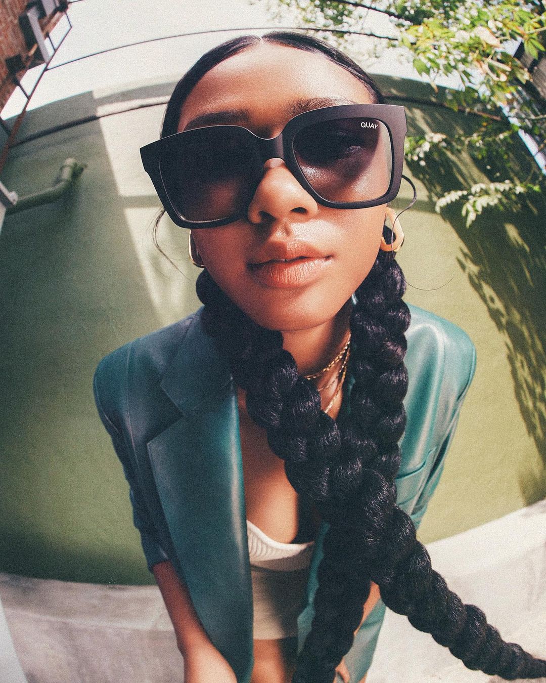 A woman with dark hair and sunglasses poses in an outdoor urban setting with a teal leather jacket and a long black braided hairstyle.